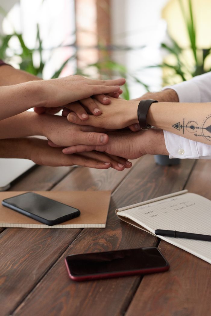 who-we-are Hands from a diverse team stack on a table symbolizing unity and teamwork in a modern office setting.